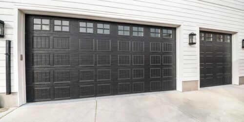 Two black garage doors with window panels and wall lighting. There is a white vinyl wood siding and a concrete driveway leading to a the three-car garage doors.