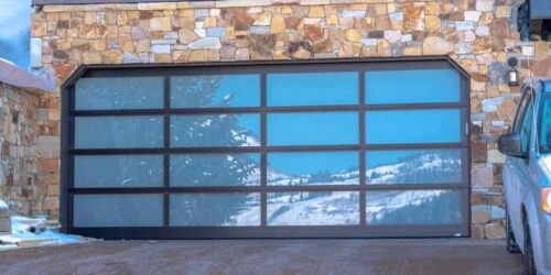 Garage door with glass panes reflecting a snowy hill landscape under blue sky. A gray vehicle is parked on the driveway of the house with stone wall.