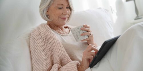 Attractive senior woman relaxing in bed with hot drink and tablet