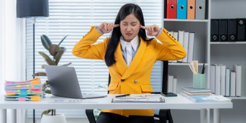 Office Noise Distraction: A woman office worker is sitting at her desk, expressing frustration by covering her ears to block out distractions, highlighting the challenges of a noisy work environment.