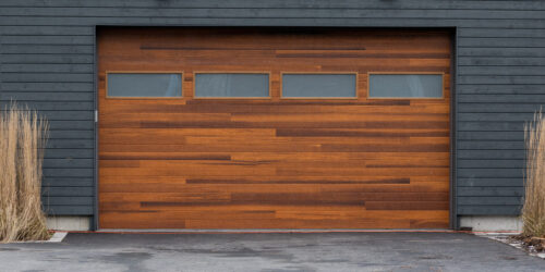 A modern brown faux wooden exterior garage door with four small horizontal glass windows. The modern door is on a luxury dark grey contemporary house with a concrete driveway.